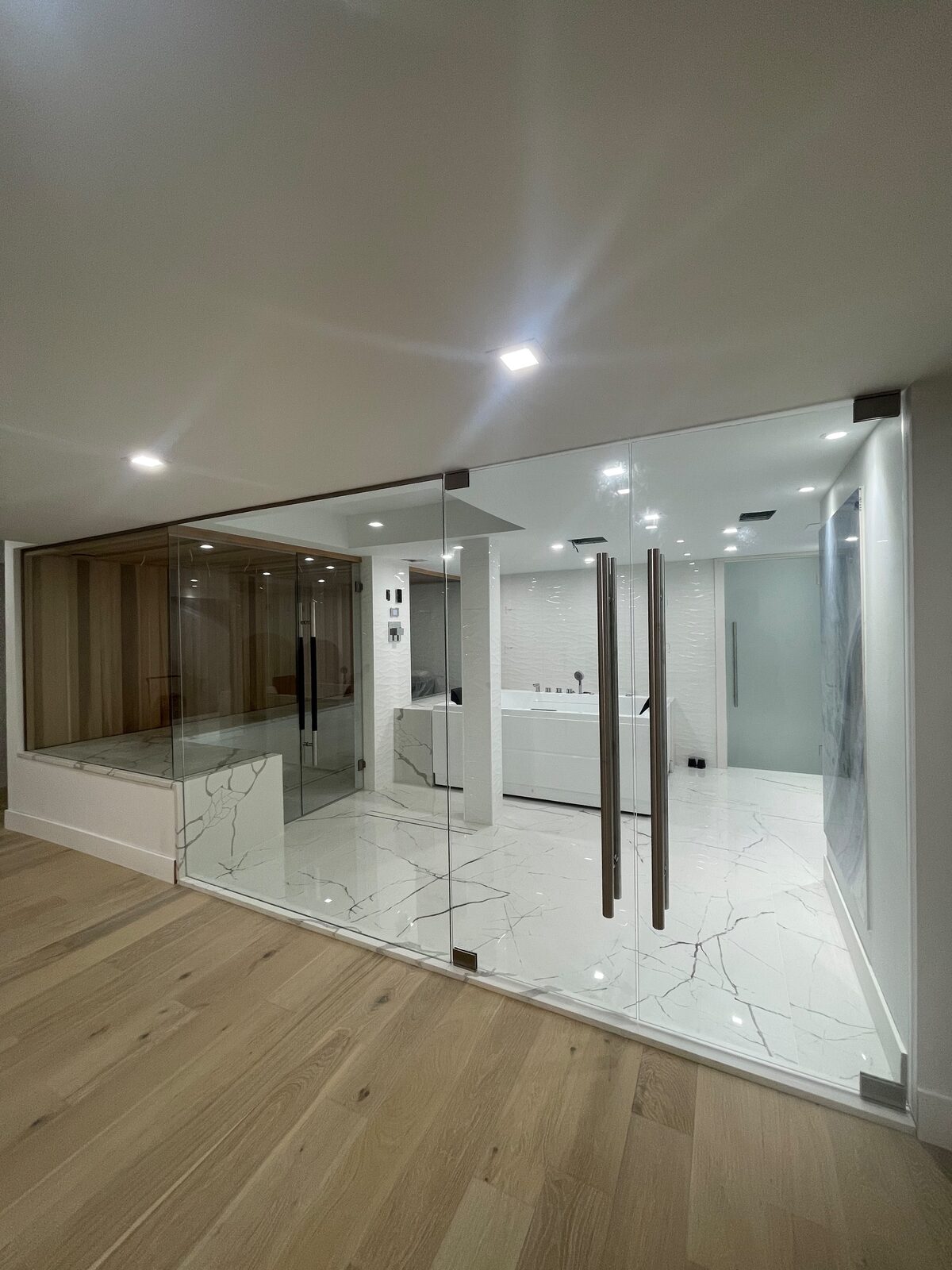 Floor-to-ceiling frameless glass wall enclosing a marble spa bathroom in a luxury Metro Detroit home